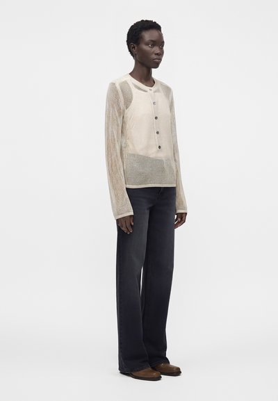 Femme portant un cardigan maille beige boutonné sur un débardeur, associé à un jean large foncé et des chaussures marron, debout sur un fond uni.