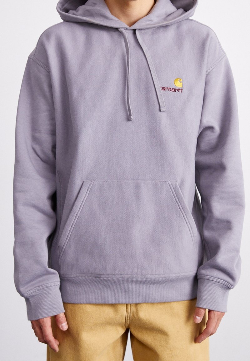 Personne portant un sweat à capuche Carhartt violet clair avec une poche avant et un pantalon jaune, debout devant un fond blanc uni.