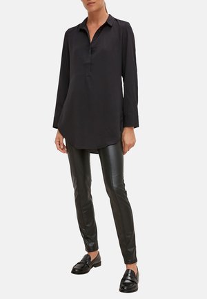 Mujer que lleva una blusa negra holgada de manga larga, pantalones pitillo de cuero negro y mocasines negros, de pie frente a un fondo liso.