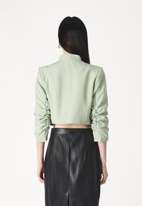 Chaqueta recortada de color verde claro con un cuello estructurado y mangas fruncidas, combinada con una falda de cuero negra de tiro alto. Textura suave.