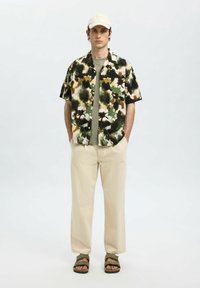 Kurzarmhemd mit einem grünen, braunen und cremefarbenen Blumenmuster, kombiniert mit einem hellgrünen T-Shirt, beigen Hosen und braunen Sandalen.