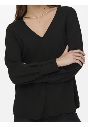 Woman wearing a long-sleeve black V-neck blouse with buttoned cuffs, crossed arms, and a simple gold necklace against a white background.