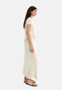 Kremowa lniana bluzka z krótkimi rękawami, zestawiona z pasującą długą spódnicą z frędzlami u dołu. Brązowe sandały z zielonymi akcentami.