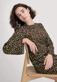 Grünes Kleid mit schwarzem Leopardenmuster, langen Glockenärmel und runden Ausschnitt. Modell sitzt auf einem Holzstuhl vor einem neutralen Hintergrund.