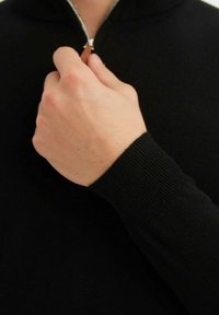 Sweat à zip noir avec des poignets côtelés et des détails de fermeture éclair. Gros plan sur une main tenant la tirette de la fermeture éclair. Texture douce, design ajusté.