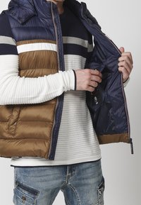 Puffer vest in brown and navy, zippered design with hood, paired with a white ribbed long-sleeve shirt. Inner pocket visible. Denim trousers.