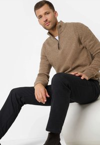 Bruine, gestructureerde ritssweater met ribgebreide boorden, gedragen over een wit shirt, gecombineerd met zwarte broek en bruine enkellaarzen.