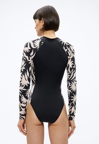 Žena s krátkymi tmavými vlasmi, oblečená v čiernych plavkách s dlhým rukávom, s béžovými kvetinovými vzormi na rukávoch a stranách, stojaca pred jednoduchým pozadím.