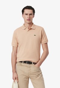 Camisa polo de melocotón clara con cuello acanalado y mangas cortas, que presenta un logo de cocodrilo bordado en verde. Combinada con pantalones beiges y un cinturón negro.