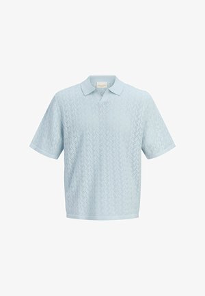 Chlapec stojaci a usmievajúci sa, oblečený v svetlomodrej textúrovanej polokošeli, béžových šortkách, bielych ponožkách a béžových mokasínach.