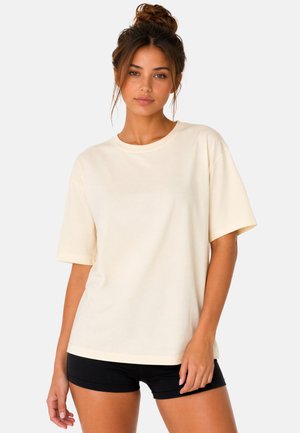 Jeune femme aux cheveux bruns en chignon portant un t-shirt oversize beige clair uni et un short noir, debout devant un fond blanc.