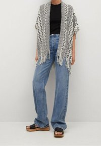 Dzianinowy szal w czarno-białe wzory z frędzlami, noszony na czarnej koszuli, zestawiony z jasnoniebieskimi szerokimi spodniami i czarnymi sandałami.