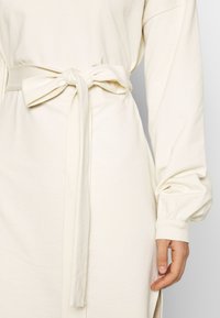 Vestido color crema con un cinturón de lazo en la cintura, mangas largas abullonadas y puños acanalados. Tejido de algodón suave con una textura lisa.