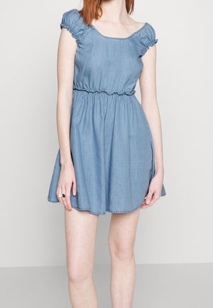 Femme portant une robe courte en denim bleu clair avec une taille froncée et des manches courtes bouffantes, debout devant un fond uni clair.