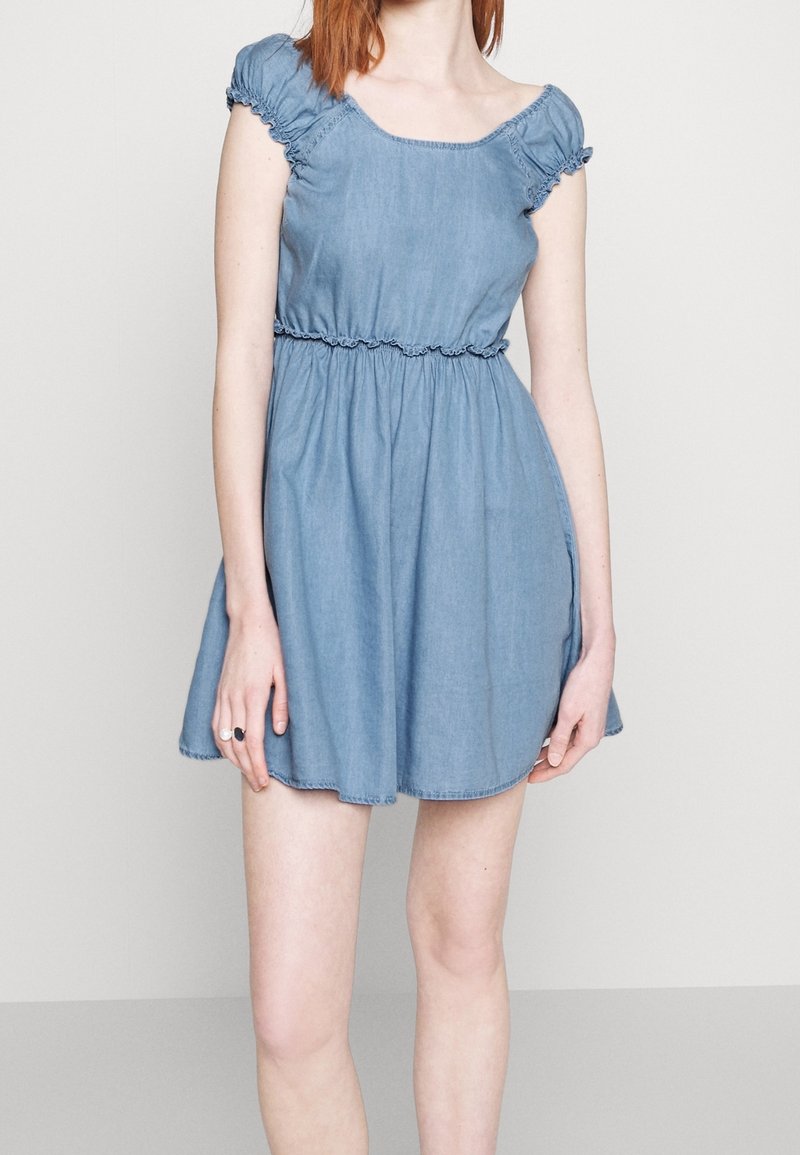 Femme portant une robe courte en denim bleu clair avec une taille froncée et des manches courtes bouffantes, debout devant un fond uni clair.