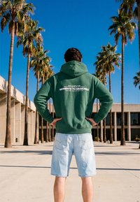 Homme debout, mains sur les hanches, portant un sweat à capuche vert avec un texte blanc et un short en jean clair, palmiers et bâtiment moderne en arrière-plan.