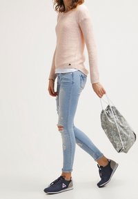Pull en tricot rose clair avec un col rond, associé à un jean déchiré bleu clair et des baskets bleu marine. Tenant un sac à cordon avec un motif.