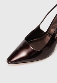 Tummanruskea lakattu nahkainen kärjellinen slingback-kengä, jossa on pehmustettu pohja valkoista taustaa vasten.