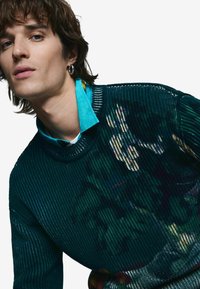 Tmavozelený rebrovaný sveter s textúrovaným pletením a kvetinovými vzormi. Navrstvený s tyrkysovou košeľou s golierom pod ním.