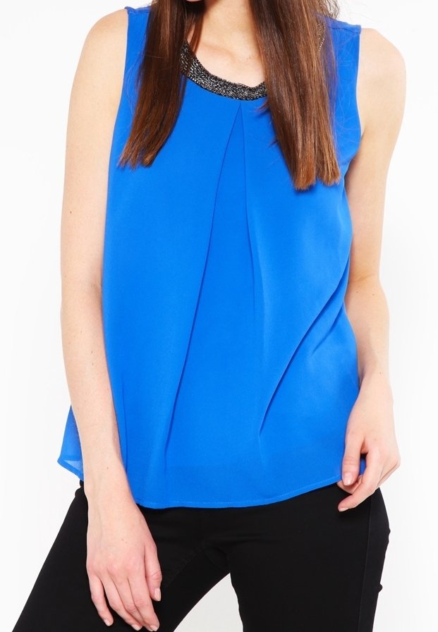 Femme portant un chemisier bleu roi sans manches avec un col orné de perles noires et un pantalon noir, debout devant un fond blanc.
