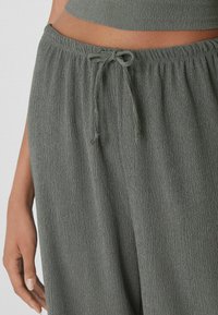 Pantalon gris texturé avec une taille élastique et un cordon de serrage, mettant en avant un tissu doux et froissé. Vue rapprochée avec une main positionnée.