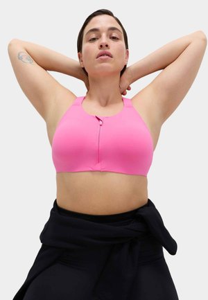 Woman with short dark hair wearing a pink sports bra and black pants, hands resting behind her head, against a plain background.