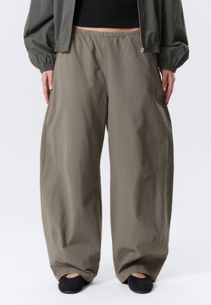 Pantalon ample vert olive porté avec des chaussures plates noires et une veste courte sombre, zippée ouverte sur un haut noir.