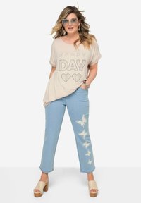 Camiseta de gran tamaño en color beige con estampado de "HAPPY DAY" en strass; jeans rectos en azul claro con diseños de mariposas blancas; zapatos de plataforma en color beige.