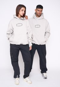 To modeller bærer matchende oversized lysegrå hættetrøjer med en sort grafik på brystet, parret med sorte bukser og sneakers.