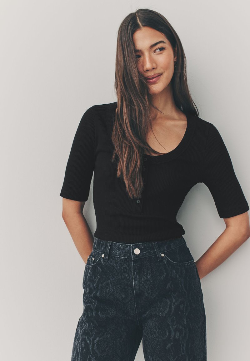 Top nero a maniche corte con motivo a coste, scollo rotondo e dettagli con bottoni, abbinato a jeans scuri a vita alta con motivo.