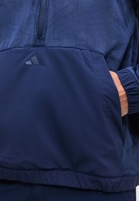 Χέρι στην τσέπη ενός ναυτικού μπλε μπουφάν με υφασμάτινη υφή στο πάνω μέρος και λείο στο κάτω, με διακριτικό λογότυπο Adidas μπροστά.