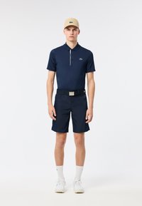 Marinblå pikétröja med en grön logotyp, tillsammans med matchande marinblå shorts. Outfiten inkluderar en beige keps och vita sneakers med texturerade sulor.
