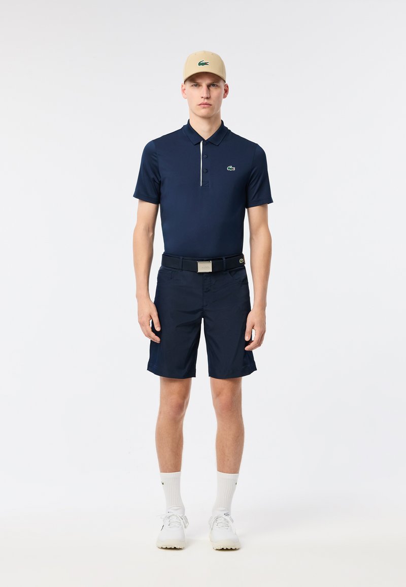 Polo shirt blu navy con logo verde, abbinato a pantaloni corti blu navy. L'outfit include un cappellino beige e sneakers bianche con suole testurizzate.