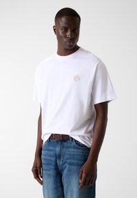 Biała bawełniana koszulka z okrągłym dekoltem i małym beżowym geometrycznym logo na lewej piersi, w połączeniu z niebieskimi jeansami.