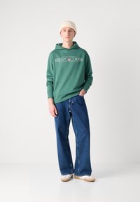 Felpa verde con testo e logo bianchi, abbinata a jeans denim blu larghi. Il modello indossa un beanie beige e scarpe chiare.