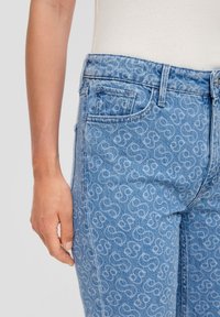 Jean en denim bleu clair avec un motif répétitif sur l'ensemble. Le design comprend des poches classiques et des détails de couture visibles.
