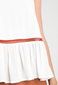 Top bianco texturizzato con orlo svasato, caratterizzato da una striscia rossa orizzontale in vita. Tessuto leggero con una leggera lucentezza.