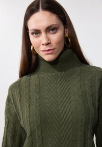 Pull tricoté vert olive à col montant, avec des motifs en câble texturés sur l'ensemble. Matière douce en mélange de laine.