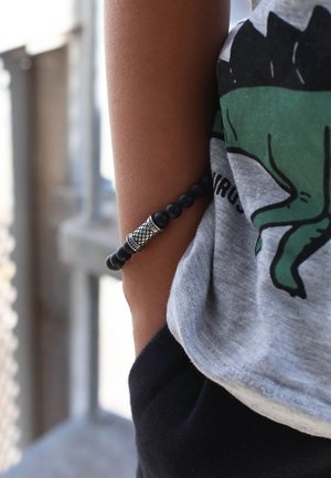 Zwarte kralenarmband met een zilveren decoratief accent, gedragen om een pols, naast een grijs t-shirt met een dinosaurustekening.