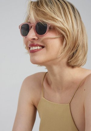 Różowe, okrągłe okulary przeciwsłoneczne z ciemnymi szkłami, z drobnymi metalowymi akcentami. Modelka ma krótkie, blond włosy i nosi jasnobrązową bluzkę.