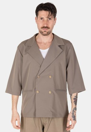 Uomo con capelli corti e scuri e avambraccio tatuato indossa una giacca taupe doppiopetto a maniche tre quarti sopra una camicia bianca.