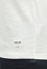 Weißes Baumwoll-T-Shirt mit strukturiertem Finish, mit einem rechteckigen Etikett auf der Rückseite, auf dem in schwarzer Schrift steht: "!SOLID DENMARK EST. 1999."