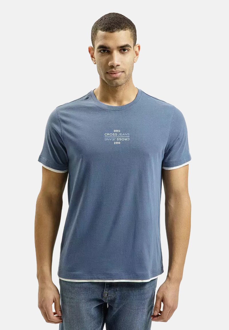 Niebieska bawełniana koszulka z okrągłym dekoltem, krótkimi rękawami i białymi akcentami. Z przodu znajduje się nadrukowany tekst "CROSS JEANS 1990" w białym kolorze.