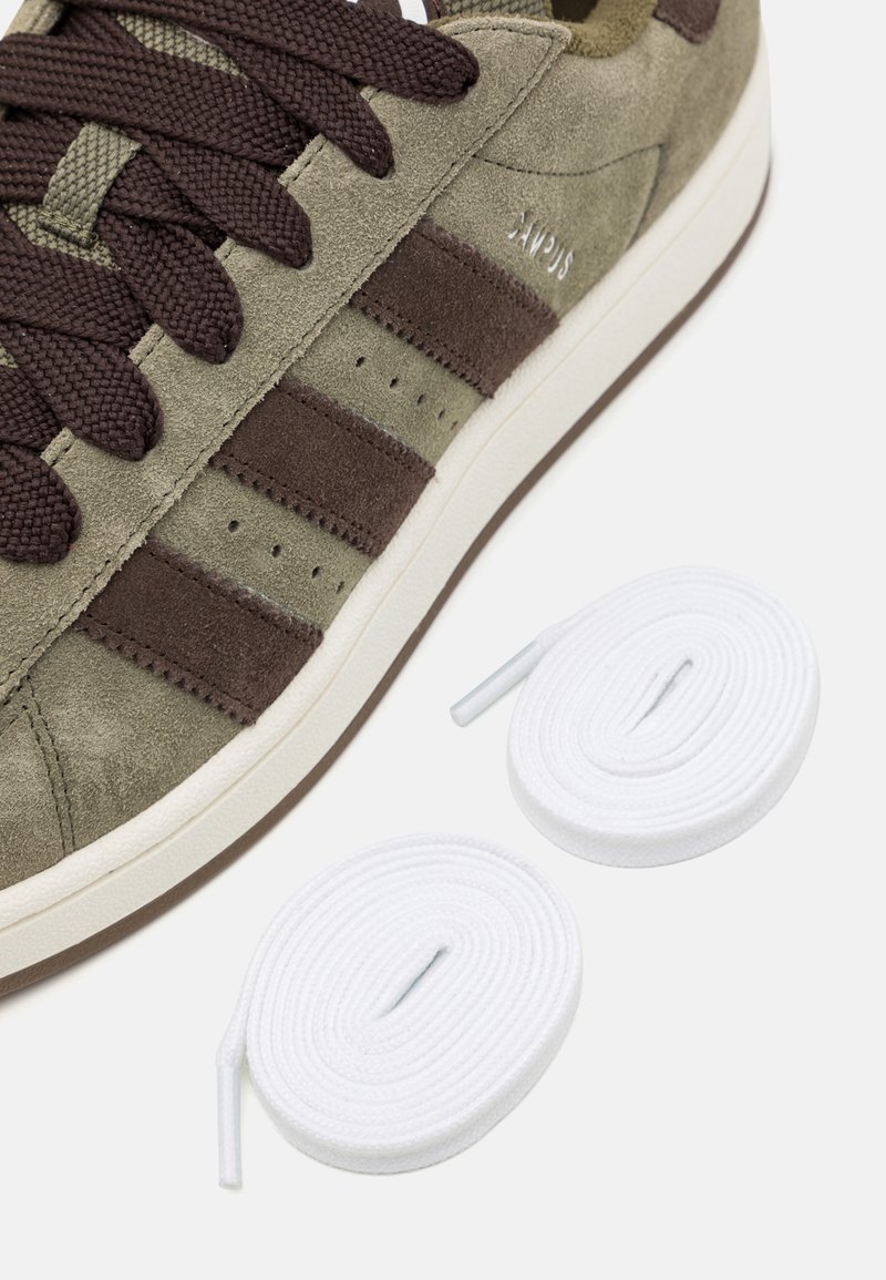 Baskets en suede vert avec des accents et des lacets marron, comportant trois rayures marron. Comprend deux lacets de rechange blancs sur un fond blanc.