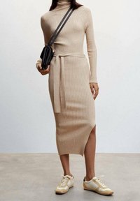 Abito a maglia beige a coste con collo alto e cintura in vita, dotato di uno spacco laterale. Abbinato a sneakers crema e gialle e una borsa nera.