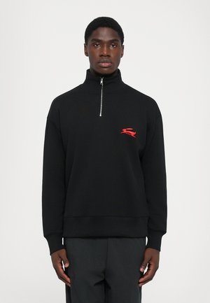 Young man wearing a black zip-up sweatshirt with a red embroidered logo on the chest, standing against a plain white background.