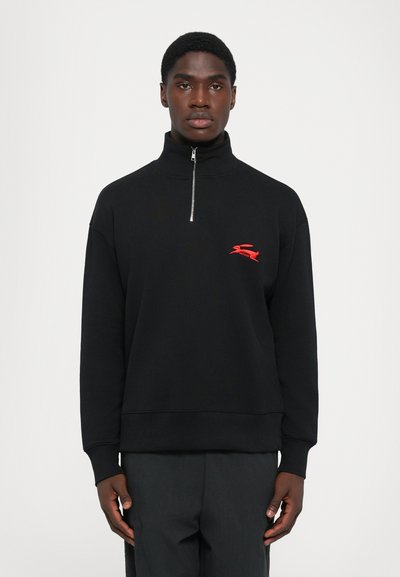 Jeune homme portant un sweat à capuche noir zippé avec un logo rouge brodé sur la poitrine, se tenant devant un fond blanc uni.