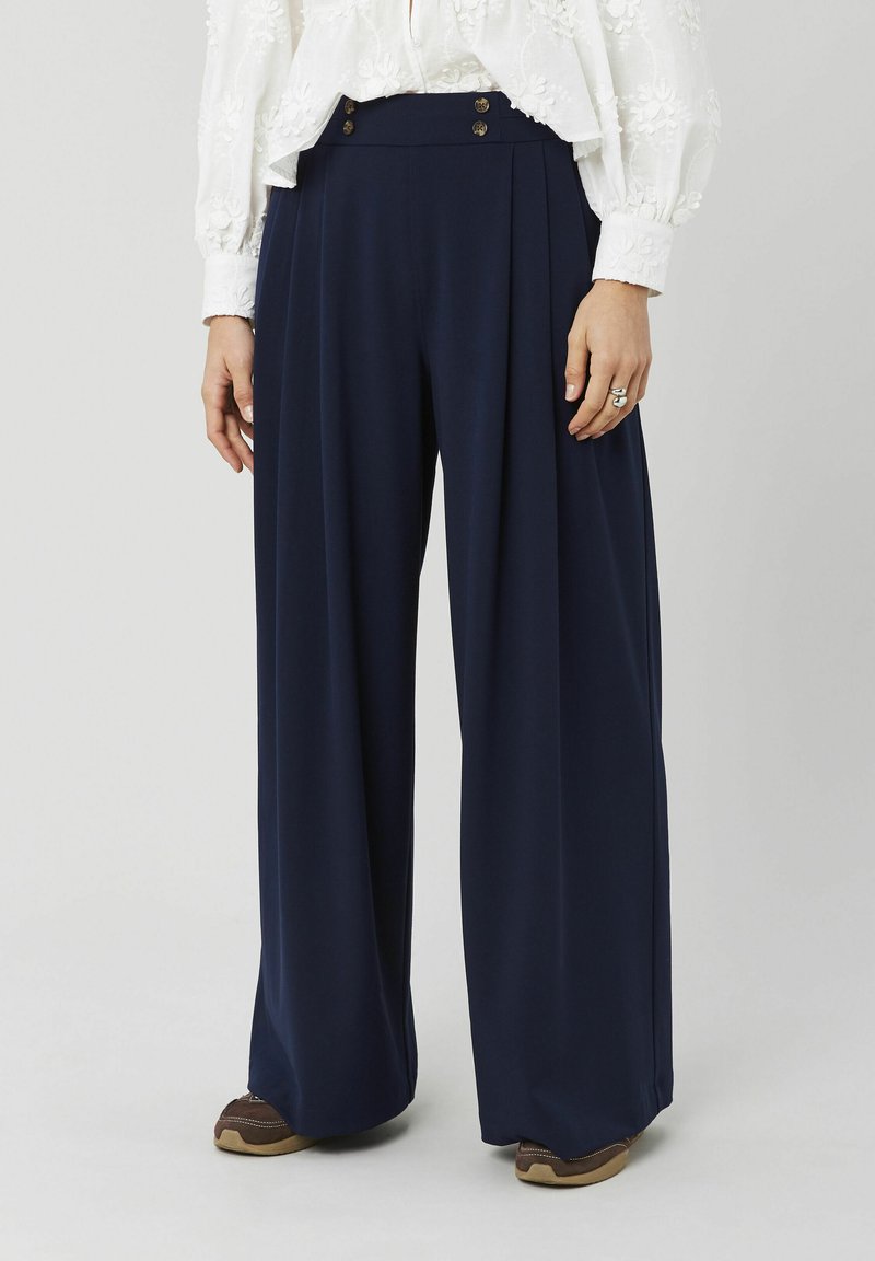 Femme portant un pantalon large taille haute bleu marine avec des détails à boutons, assorti à des chaussures marron et un chemisier blanc brodé.