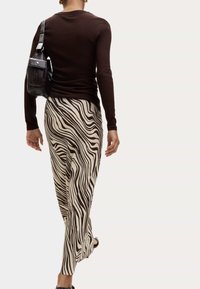 Hnědý přiléhavý svetr, béžové kalhoty s černými vlnitými pruhy a černá kabelka nošená přes tělo. Hladká textura a slim fit.