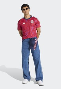 T-shirt de sport rouge avec un motif floral rose et des accents bleu marine, associé à un jean bleu et une casquette bleu marine affichant "Adidas" en rose.